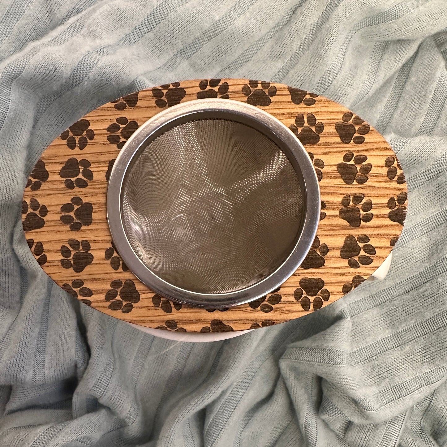 Wooden tea infuser with stainless steel basket engraved with paw prints on a light blue blanket background.