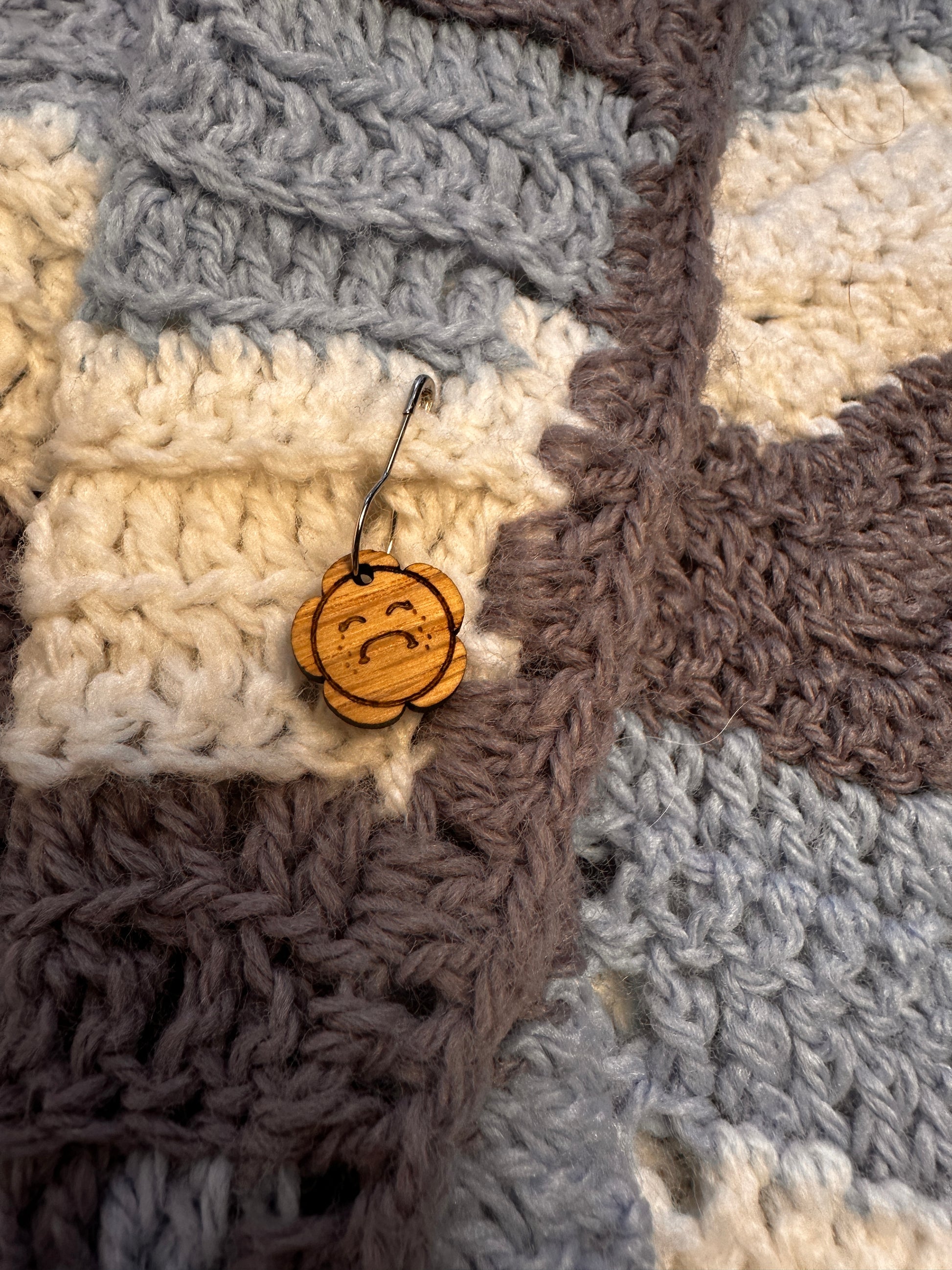 Wooden sad face flower oops stitch marker pinned to a crocheted blanket. 