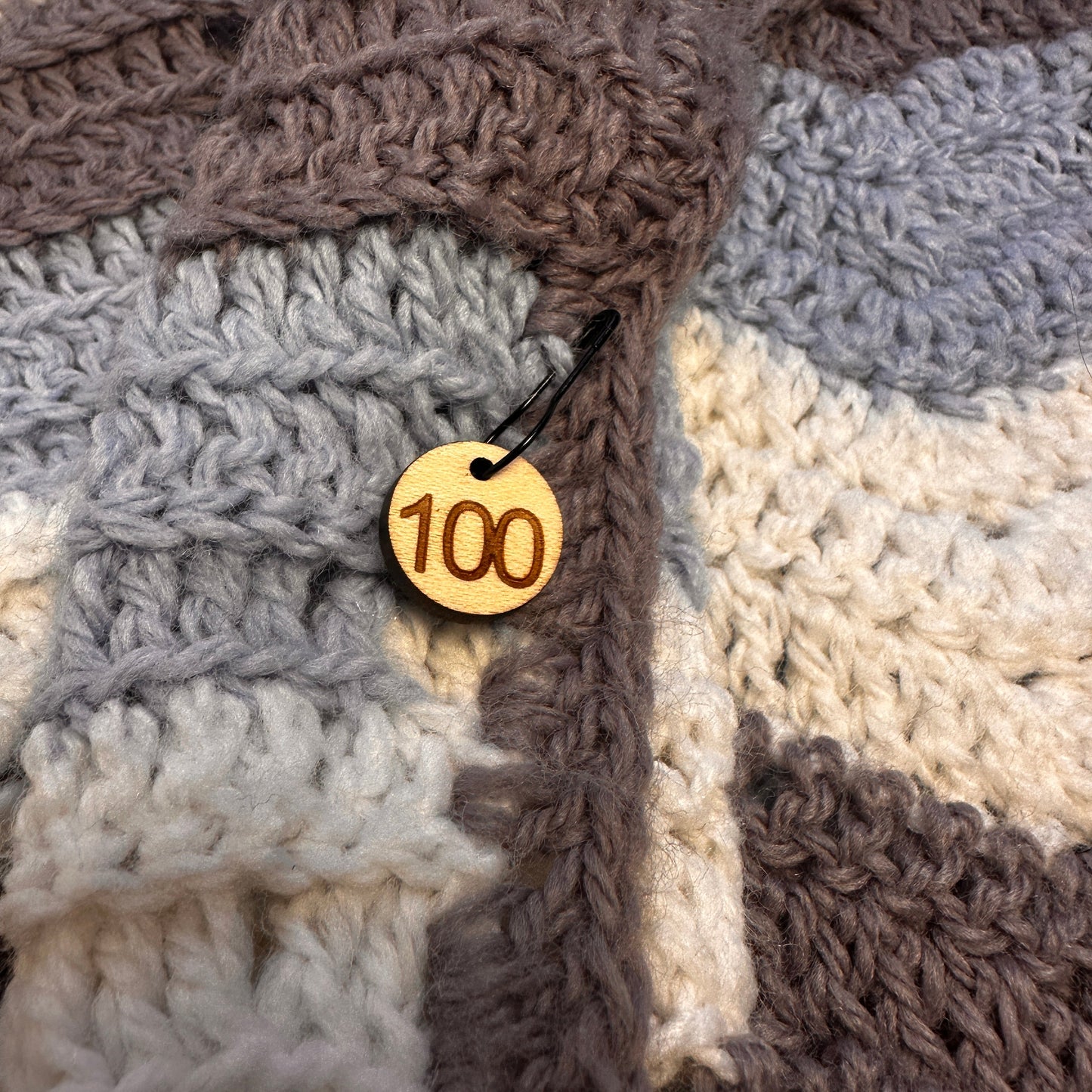 Close-up of a wooden stitch marker with '100' engraved attached to a crochet blanket. 