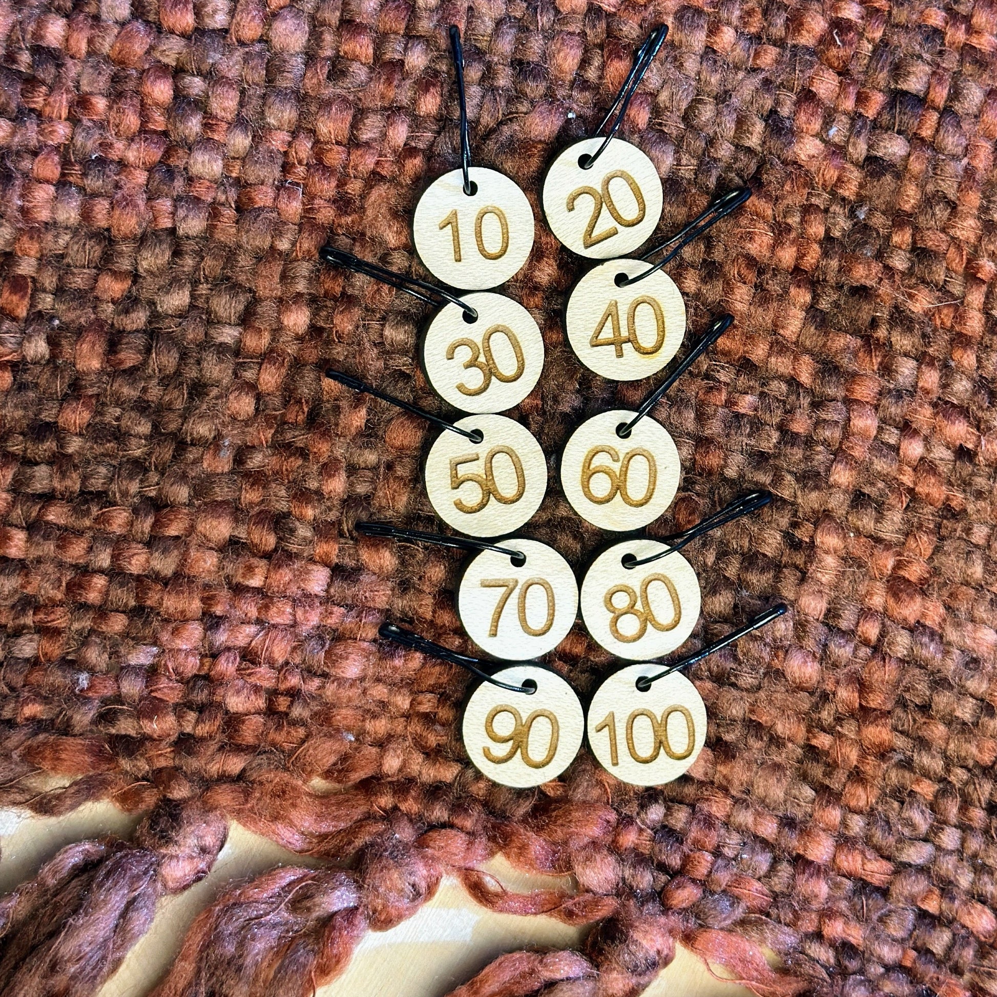 Wooden stitch marker set for rows 10-100 laying on top of an orange and rust knitted blanket. 